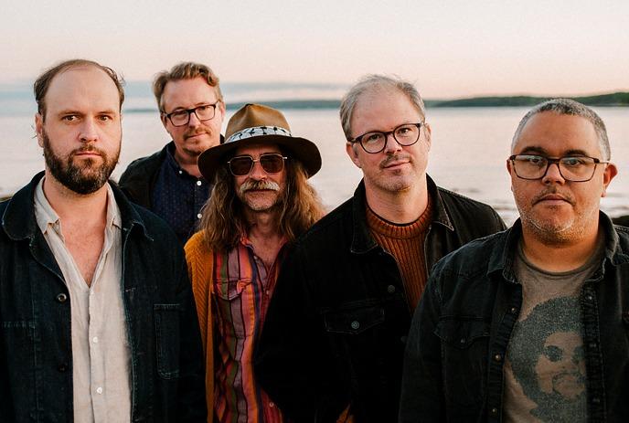 The 5 band members of Wintersleep posing for the camera in front of a body of water at sunset.