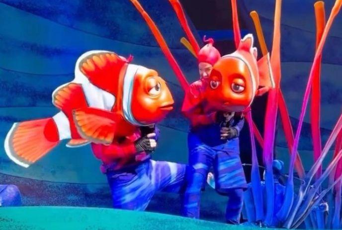 Two performers in blue costumes animate large clownfish puppets on stage, set against vibrant coral backdrop.