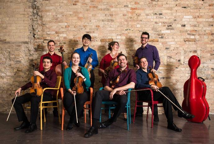 Eight artists with violins, violas, and cello posing under a bricked wall.