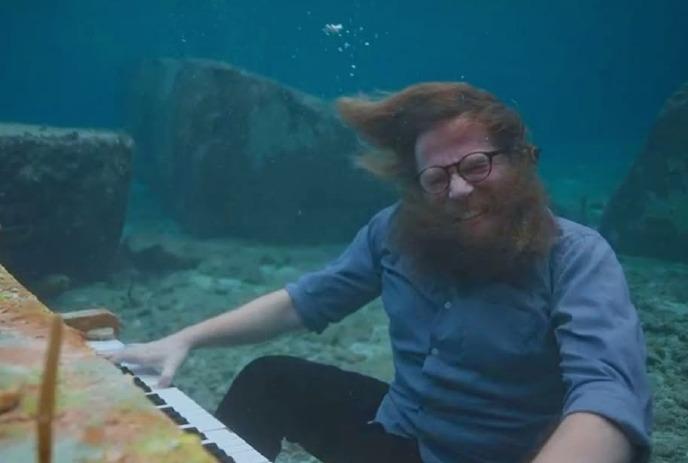 Ben Caplan plays a piano underwater with his hair floating and bubbles rising around him.