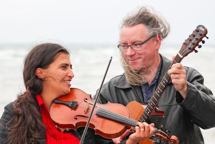Steafan Hannigan & Saskia Tomkins standing outside on a windy day, holding a violin and a guitar.