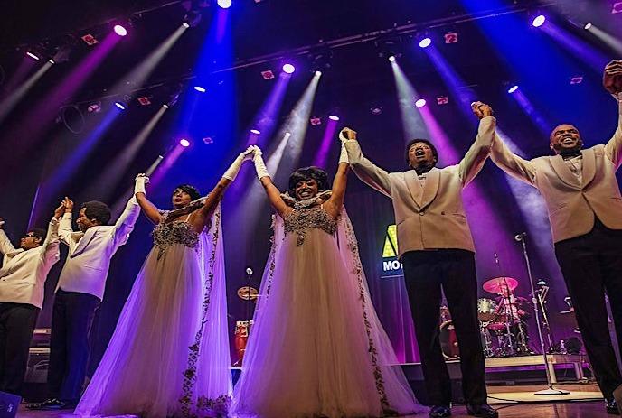 Performers in gowns and tuxedos raise arms on a lit stage with a band.
