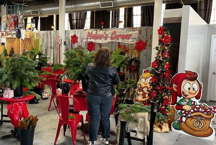 Person crafting with pine at Maker’s Corner, surrounded by red chairs, elf cutouts, and festive supplies.
