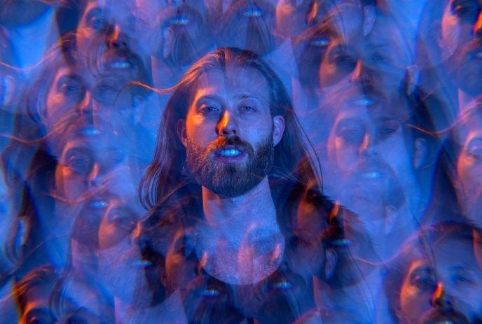 Artist with long hair and beard surrounded by kaleidoscope of blurred reflections in blue-purple light.