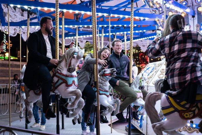 People ride carousel horses at night, smiling amid bright lights and festive energy.