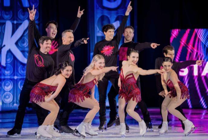 Ten skaters pose on ice in red and black costumes, smiling under blue and purple lights.