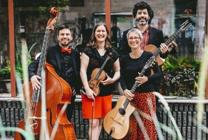 Four musicians pose with instruments outdoors by windows, smiling for a picture.