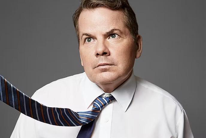 A black and white photo of Bruce McCulloch wearing a tie being blow upwards, behind a purple background.