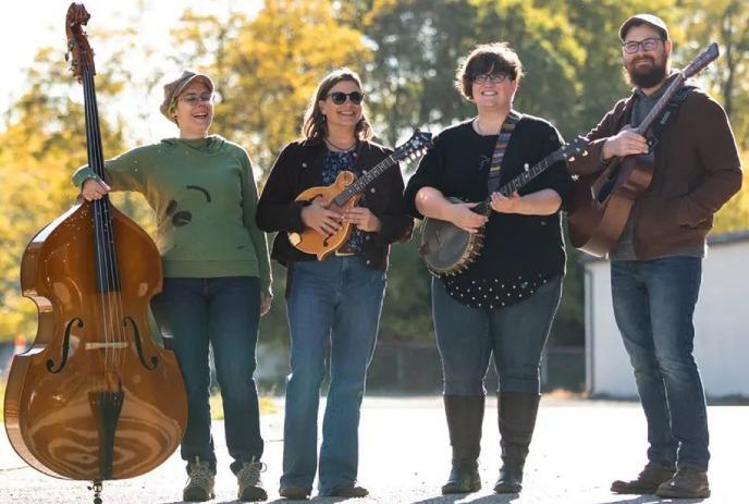 Four musicians stand outdoors with double bass, mandolin, banjo, and guitar; autumn trees and white building behind.