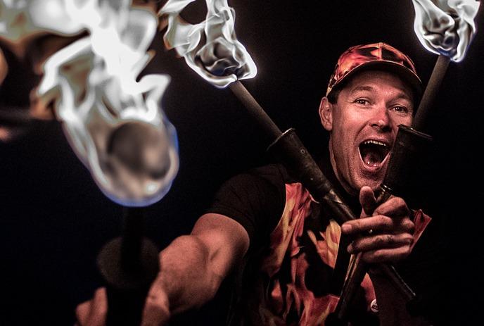 Artist holds three flaming torches in dramatic pose; dark background highlights fire and intense energy.