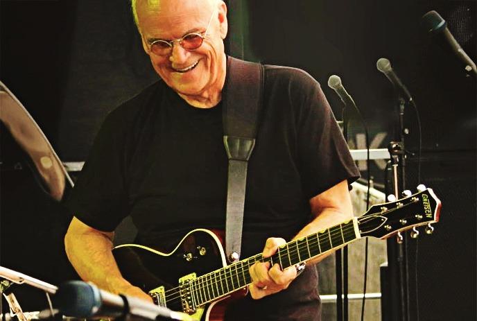 Ian Thomas performing on stage with his guitar, smiling, with his band performing with him in the background.