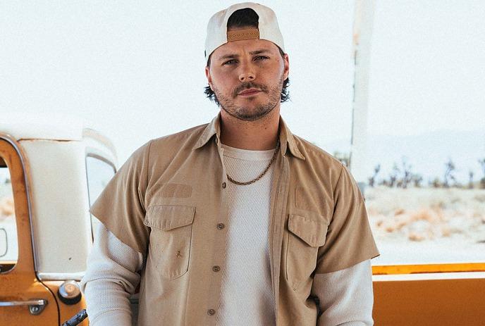 Josh Ross standing by an old orange truck in the desert, wearing a beige shirt and white cap.