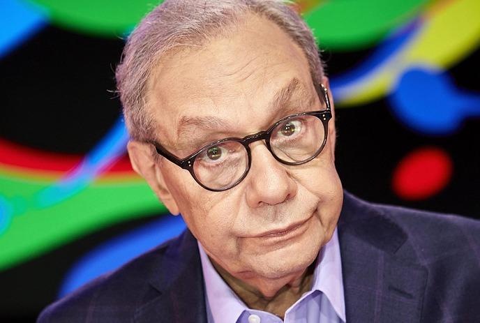 Lewis Black looking at the camera with a blank expression, wearing glasses and a suit in front of a dark background.