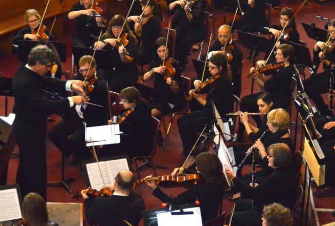 Large orchestra performing indoors; musicians in formal black attire play strings and woodwinds.