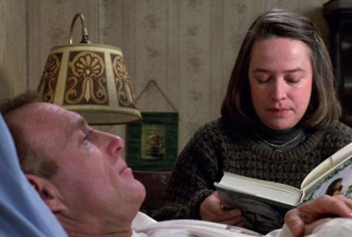 A scene from the film Misery where an individual is lying on the bed and a lady on the side reading a book.