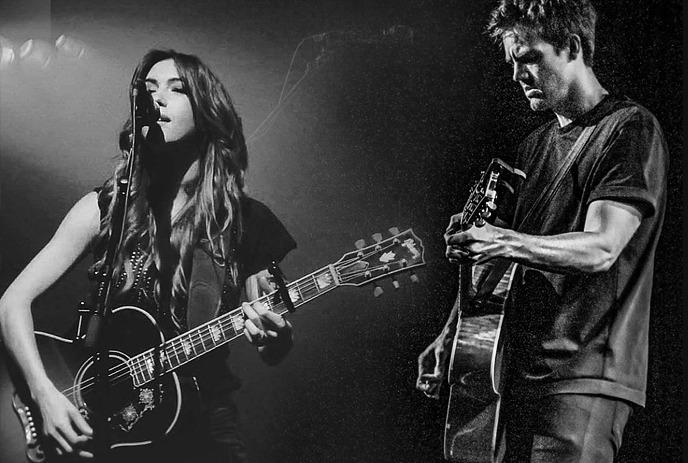 A black and white photo of Tyler Hilton & Kate Voegele on stage playing their guitars and singing.