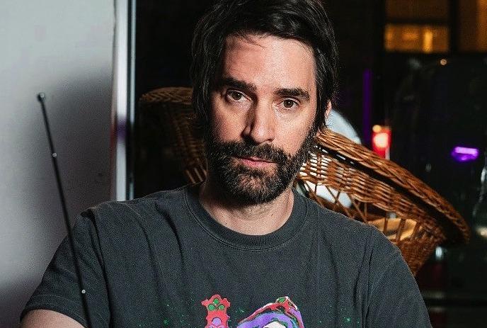 Phil Hanley looking at the camera with a blank expression in a room, sitting down.