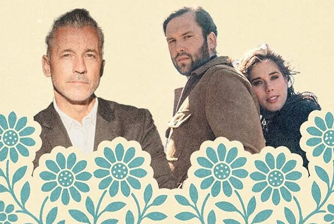 Three people stand behind blue floral border, one in suit, two in casual wear against plain beige backdrop.