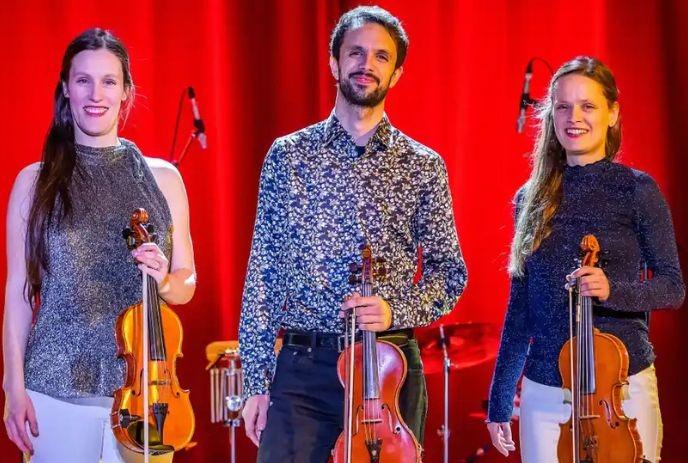 Three violinists in sparkly outfits pose on stage, instruments in hand, ready to perform under red curtains.