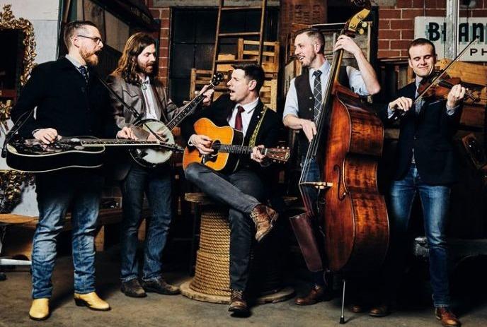 Five musicians play dobro, banjo, guitar, bass, and fiddle in rustic indoor setting with warm vintage decor.