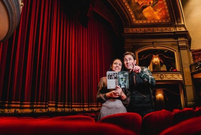 Two people stand in ornate theater before red curtain, holding program and pointing; red seats and arches visible.