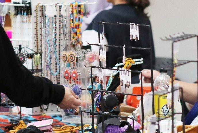 Handmade jewelry on display at market, person browsing items, vendor seated nearby with festive vibe.