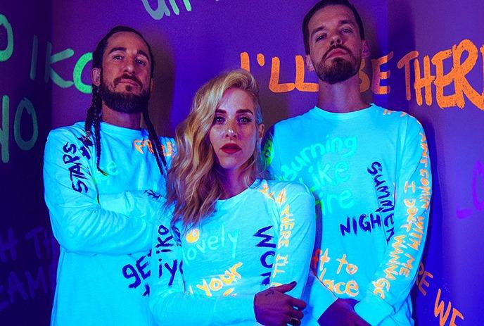 Walk off the Earth members posing in glowing white outfits with colorful text against a vibrant, graffiti-covered purple wall.