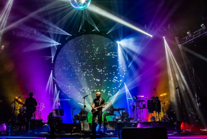 Group singing onstage with numerous lights behind them.