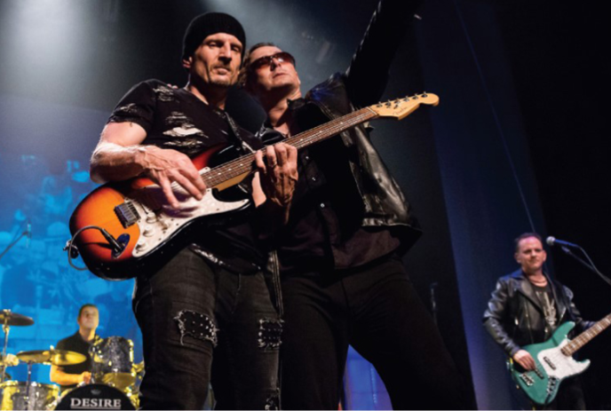 Two band members playing guitar, one member playing drums and one singing on stage.