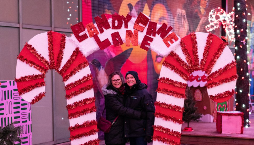 An older couple poses under a sign that says Candy Cane Lane with illuminated candy canes on either side.