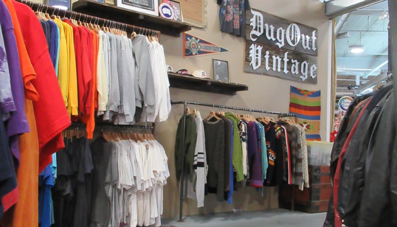  A vintage shop with clothing racks filled with sweaters. A sign reads DugOut Vintage.