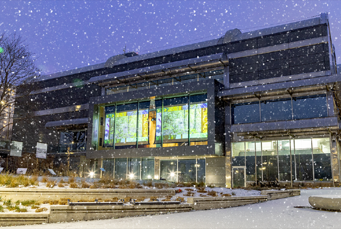 Snow falls on Museum London with glowing stained glass, lighting up steps and planters in winter dusk.