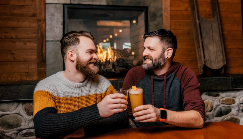 Two men clink glasses in front of a fireplace in a cozy brewery.